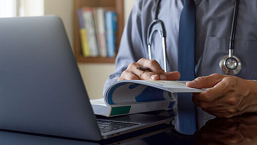 Arzt arbeitet am Laptop und liest ein Buch | Foto: ©NIKCOA – stock.adobe.com